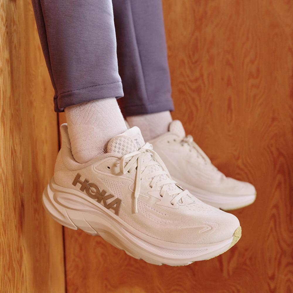 Beige Hoka running shoes worn with gray pants on a wooden floor.