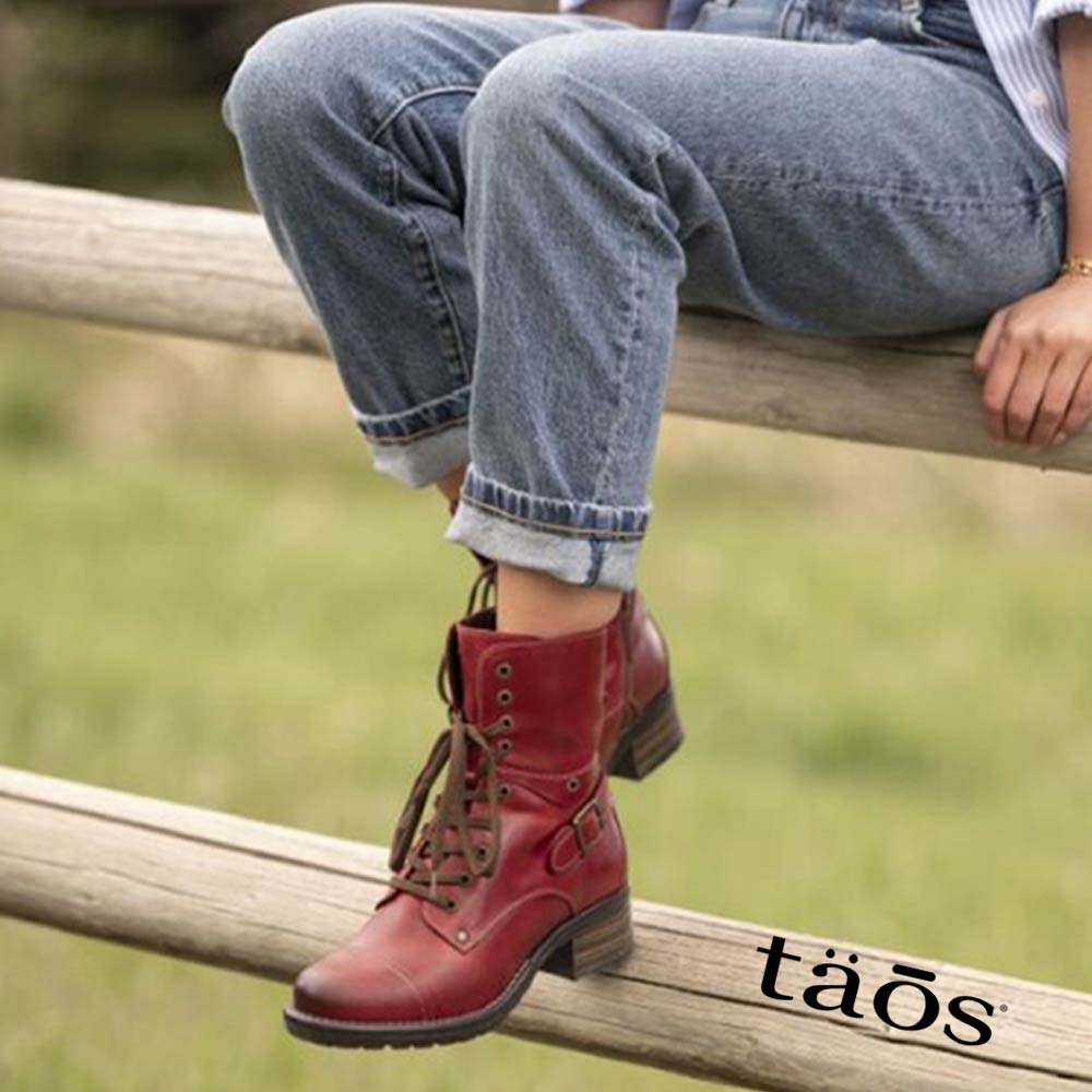 Person wearing red boots and blue jeans sitting on a wooden fence with 'taos' branding.