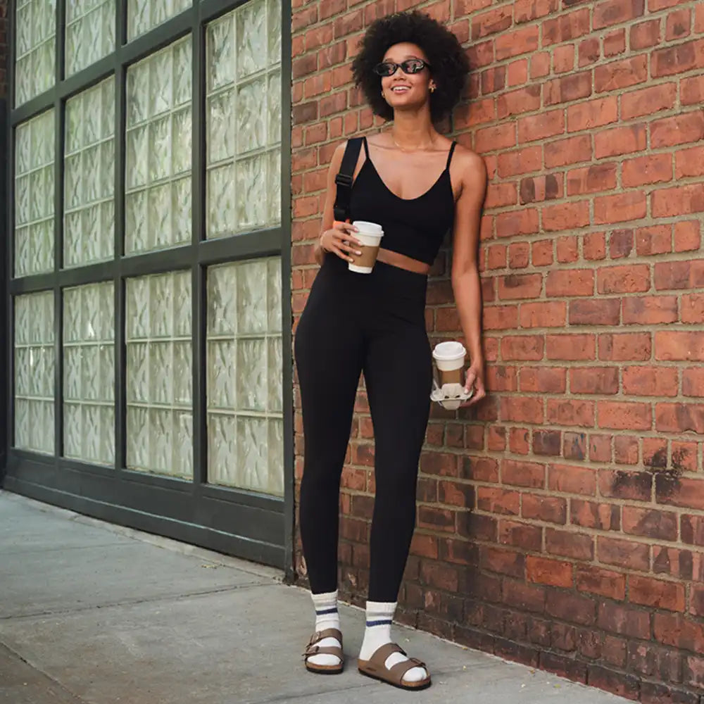 Woman in black athletic wear holding coffee cups against a brick wall.